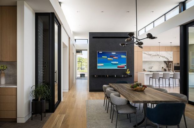 A bright, contemporary open-plan dining and living area with a long timber dining table, upholstered dining chairs, a large neutral rug, and sculptural black pendant lighting. A dark feature wall with a mounted artwork anchors the dining zone, while a warm timber kitchen with a large marble-look splashback and island is visible beyond. Full-height glazing and clerestory windows bring in abundant natural light, and a corridor and glazed interior door are visible to the left.