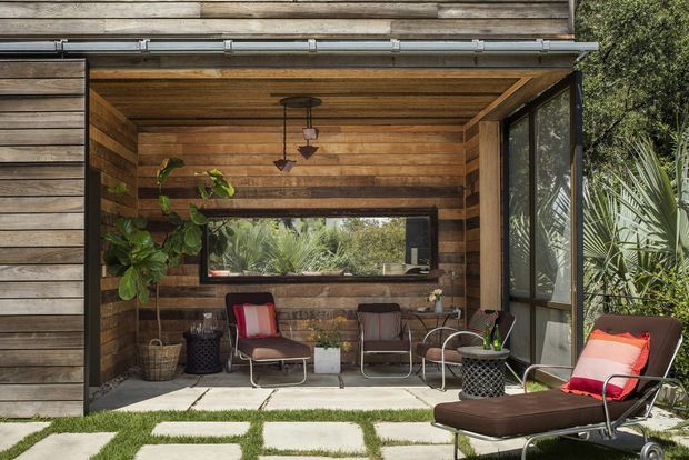 A small, enclosed outdoor living pavilion clad in horizontal timber, with a timber-lined ceiling, large glazed openings, and a concrete-paver floor separated by strips of grass. The space contains relaxed lounge seating, a long low sun lounger, potted plants, and pendant lighting, creating a warm indoor-outdoor retreat surrounded by dense planting.