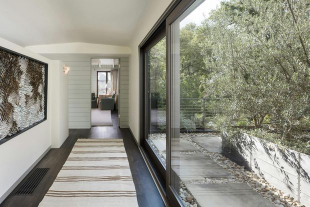 A bright, contemporary interior hallway or landing with dark timber flooring, a long striped runner, white walls and horizontal wall panelling. Full-height glazing along one side overlooks a planted, stepped outdoor area, while a large framed textured artwork forms a feature on the opposite wall. An open doorway leads toward a furnished room beyond.
