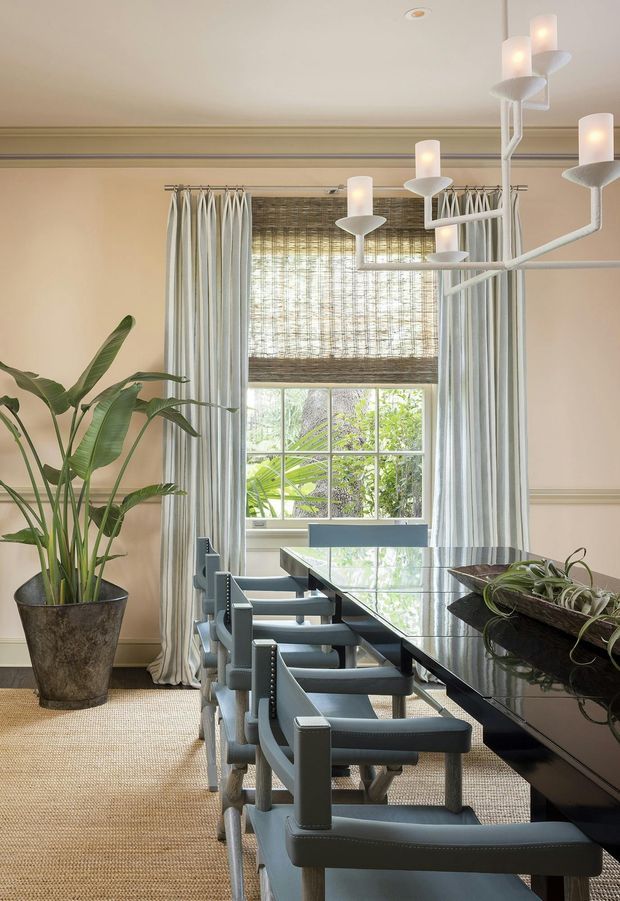Elegant dining room with a long glossy dark dining table, multiple upholstered dining chairs, full-height striped curtains, a woven Roman blind, and a sculptural white multi-arm chandelier. The room has warm beige walls with traditional picture-rail and dado-rail mouldings, a textured natural-fibre carpet, and a large potted tropical plant beside the window.