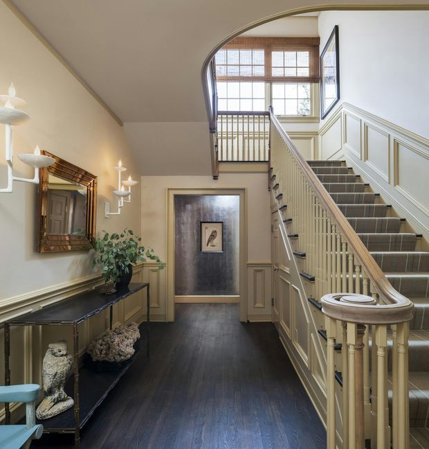 A refined traditional residential stair hall with a dark timber floor, cream painted walls, extensive raised wall panelling, and a carpeted staircase with turned balusters and a curved handrail. The stair rises to a landing framed by a large arched opening and timber multi-pane windows. A slim console table, ornate mirror, wall sconces, and decorative objects line the hallway.