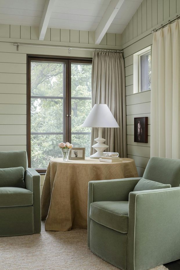 A calm, light-filled sitting room with pale green horizontal wall panelling, exposed white ceiling beams, and large dark timber-framed glazed doors overlooking leafy vegetation. Two sage-green upholstered armchairs face a small round table draped in natural beige fabric, styled with a sculptural white lamp, flowers, books, and framed photographs. Full-length cream curtains and a woven neutral rug complete the softly layered interior.