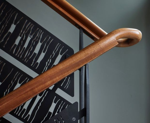Close-up architectural detail of a sculptural timber handrail with a smoothly curved end, mounted beside a dark perforated metal balustrade. The composition emphasizes the craftsmanship, warm wood grain, black metalwork, and cool grey wall surface.