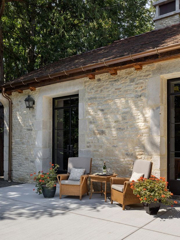 A sunlit exterior patio beside a traditional stone building. The facade features pale rough-cut masonry, dark-framed glazed French doors and windows, exposed timber eaves, copper-coloured gutters, and a shingle-clad pitched roof. Wicker lounge chairs and a small table create an outdoor seating area, with potted flowering plants on a light concrete terrace.