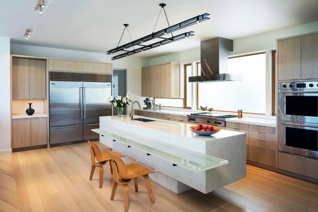 Bright, spacious contemporary kitchen with a large white stone island, integrated seating, extensive light timber cabinetry, stainless-steel appliances, and a prominent sculptural rangehood. Linear black pendant lighting contrasts with the pale finishes, while timber flooring and broad windows create a warm, airy atmosphere.