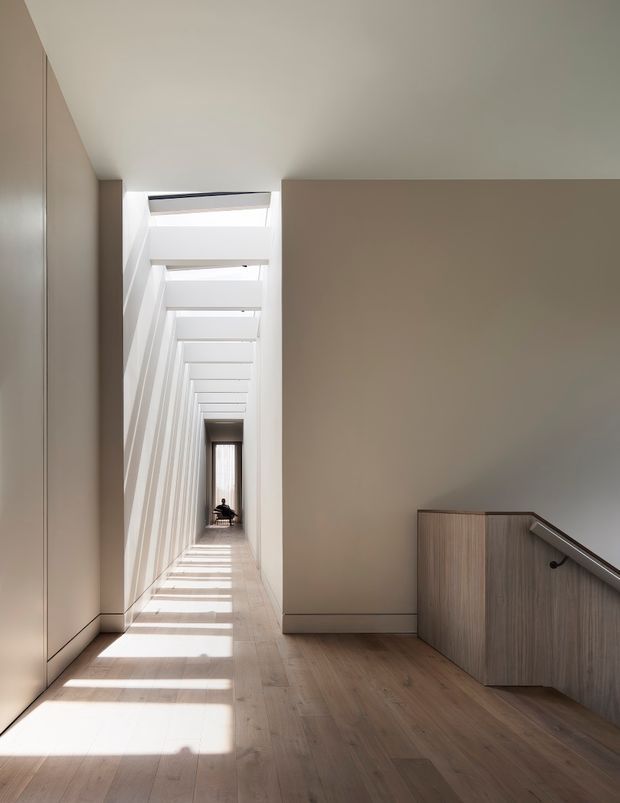 A minimalist interior hallway or landing with a long, narrow corridor illuminated by repeated linear skylights. Pale taupe walls, light timber flooring, and flush wall surfaces create a calm, highly ordered composition. A timber-clad stair balustrade and dark handrail occupy the foreground, while strong bands of sunlight extend down the corridor toward a distant doorway or figure.