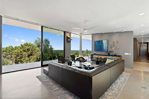 Bright contemporary living room with a large dark leather sectional arranged around a glass coffee table and rug. Full-height black-framed glazing opens the room visually to a leafy landscape, while a grey feature wall holds a wall-mounted television, artwork and low media console. A ceiling fan, recessed lighting and a hallway extending to the rear complete the open-plan interior.