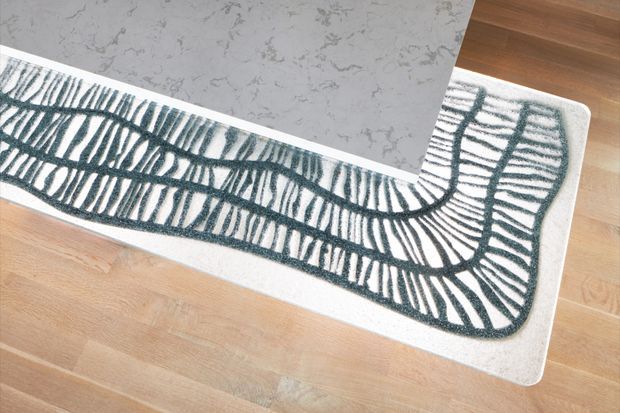 Top-down view of a curved kitchen island or benchtop with a pale grey stone-look surface, set over light timber flooring. A long, rounded rectangular area rug in white with a dark charcoal organic linear pattern follows the island’s contour.