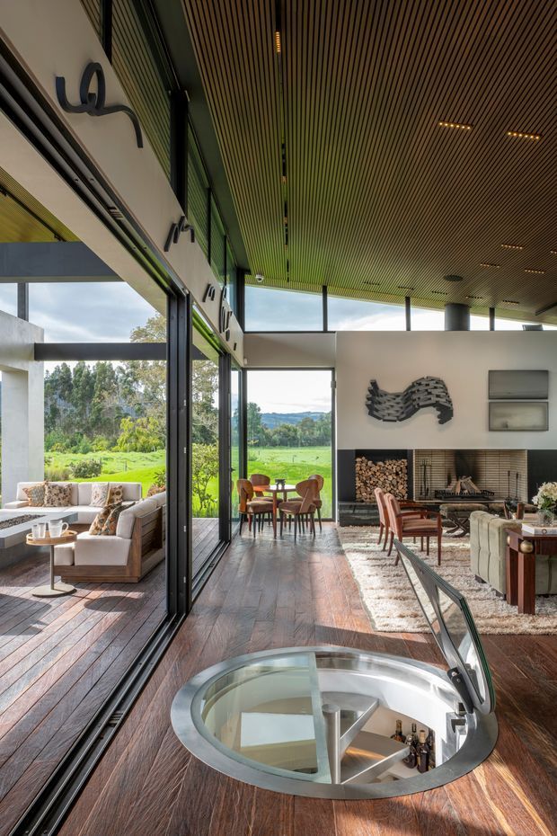 A spacious, refined open-plan living and dining area with floor-to-ceiling black-framed glazing overlooking a green landscape. The room features dark timber flooring, a linear slatted timber ceiling, a sculptural fireplace with integrated firewood storage, contemporary seating, and a circular glazed stair opening in the foreground. Strong indoor-outdoor connection and abundant natural light define the space.