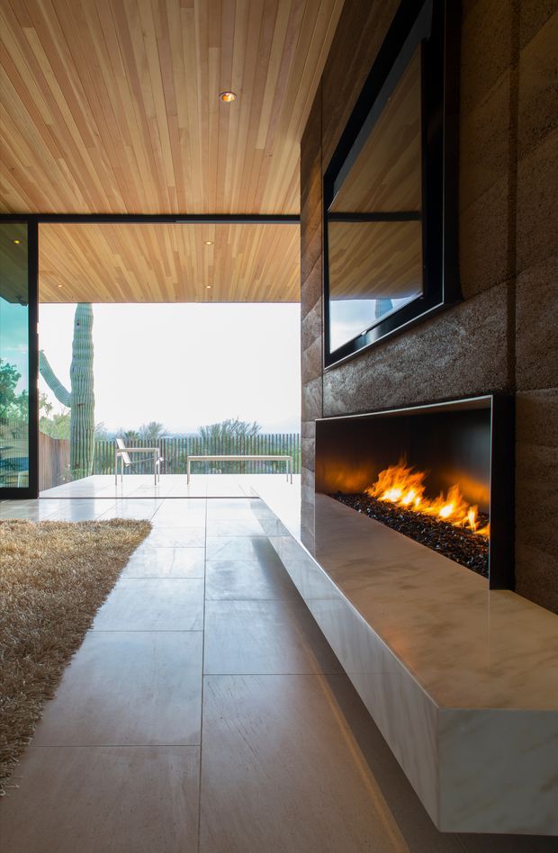 A contemporary interior lounge or living area opening onto an outdoor terrace. The room features a full-height black-framed glazed opening, a warm timber-lined ceiling, large pale stone or porcelain floor tiles, and a dramatic linear built-in fireplace set within a textured brown wall. A minimalist outdoor seating area and cactus landscape are visible beyond the glazing.