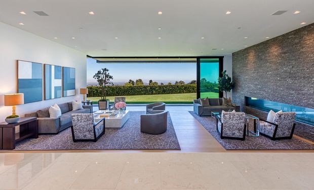 A spacious, high-end contemporary living room with a large sectional sofa, sculptural lounge chairs, coffee table, patterned area rug, and a long linear fireplace set into a dark textured stone feature wall. Full-height glazing opens the room toward a landscaped terrace and distant ocean or horizon views. The room has a polished pale stone floor, white ceiling with recessed lighting, and a refined neutral palette with blue and grey accents.
