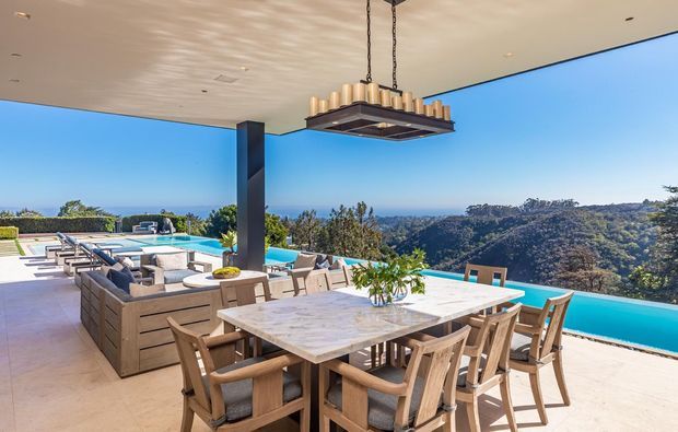 A spacious covered outdoor dining and entertaining area overlooking a swimming pool and expansive hillside landscape. The space features a large marble-look dining table with timber dining chairs, a timber outdoor lounge setting, pale tiled flooring, a dark structural column, and a dramatic suspended linear pendant light. Floor-to-ceiling open sides create a seamless connection to the pool and panoramic view.