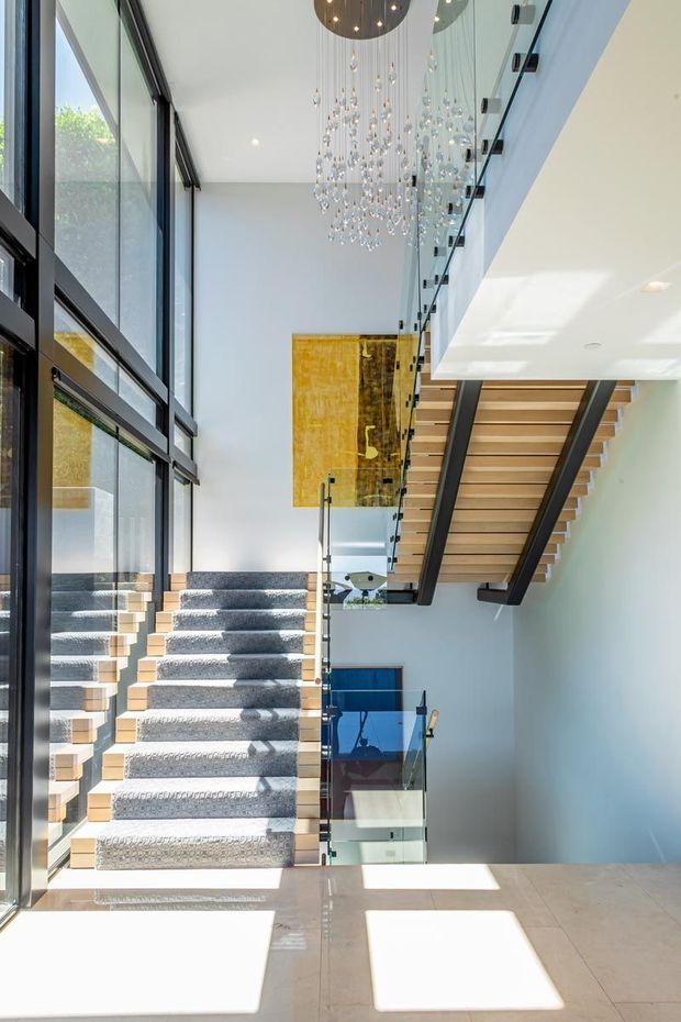 Bright contemporary stair hall with a sculptural split-level staircase, pale carpeted steps, timber treads, dark metal stringers, and clear glass balustrades. Full-height black-framed glazing floods the space with sunlight, while a dramatic cascading pendant hangs through the double-height void. A large gold-toned feature artwork and glimpses of lower-level living areas add visual interest.