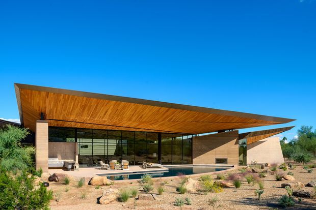 Exterior view of a low, single-storey contemporary residence in a desert landscape. The building features a dramatic broad cantilevered roof with a warm timber soffit, extensive floor-to-ceiling glazing, stone or rendered masonry walls, and an integrated outdoor terrace with a long narrow reflecting or swimming pool. Xeriscaped gravel planting surrounds the house beneath a clear blue sky.