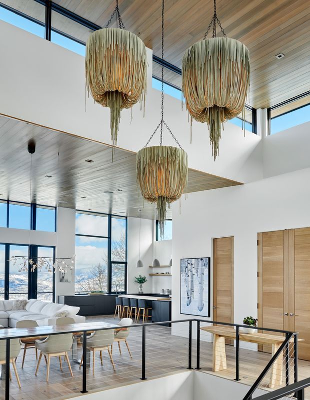 A bright, expansive open-plan interior with a double-height void, large black-framed windows, light timber ceiling, and pale tiled flooring. The space combines a living and dining area with a compact kitchen and island, connected by a black metal stair balustrade. Three oversized sculptural pendant lights with cascading natural-fibre strands dominate the ceiling. Light timber doors, white walls, and views through the glazing create an airy contemporary atmosphere.