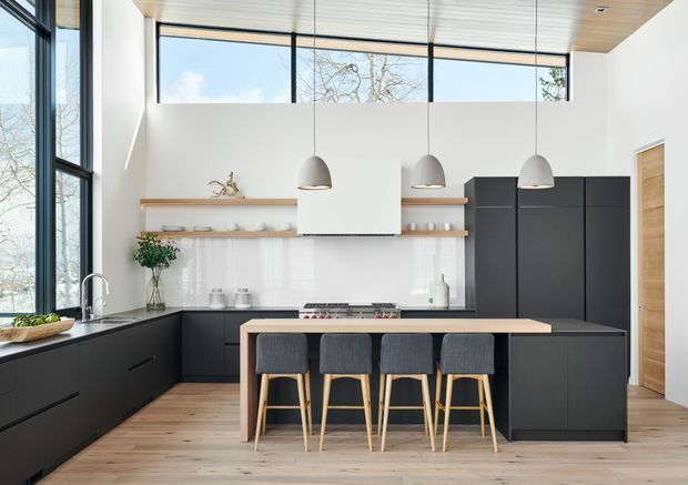 Bright, spacious contemporary kitchen with a large central island and four upholstered timber stools. The room features matte black cabinetry, a pale stone or composite island benchtop, white walls and splashback, open timber shelving, pendant lights, extensive black-framed glazing, and a light timber slatted ceiling. Light timber flooring and abundant natural light create an airy, refined appearance.