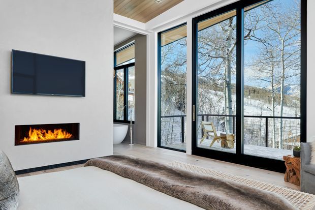 A contemporary living room with a wall-mounted flat-screen TV above a linear fireplace, expansive black-framed floor-to-ceiling glazing, and a timber-lined ceiling. A large bed-like or low upholstered seating surface occupies the foreground, while snowy trees and mountain terrain are visible outside. The room has a refined alpine outlook with warm timber and neutral finishes.