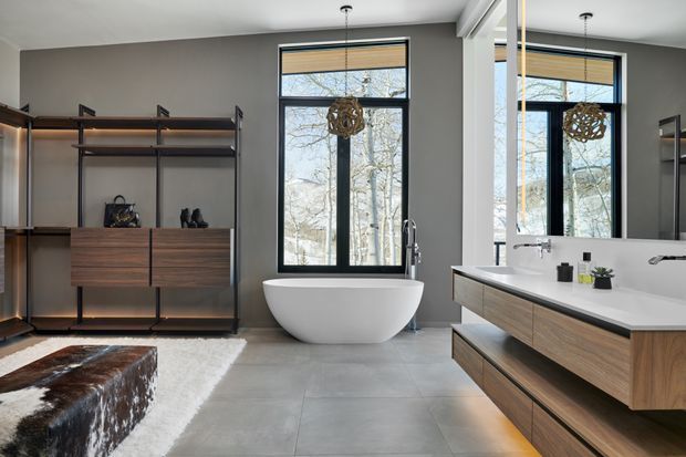 A spacious contemporary bathroom with a freestanding white oval bathtub positioned beneath tall black-framed windows. A long floating timber vanity with a white integrated basin occupies the right side, while open timber-and-black-metal shelving and cabinetry line the left wall. Large grey floor tiles, grey walls, warm concealed lighting, and sculptural pendant lights create a refined modern interior. A cowhide-covered bench and rug are visible in the foreground.