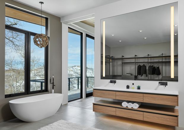 Contemporary master bathroom with a freestanding white bathtub beside expansive black-framed glazing, a large illuminated mirror, and a floating timber double vanity with open shelving. The space has light grey tiled flooring, white walls, recessed ceiling lighting, and views toward a wooded, mountainous landscape.