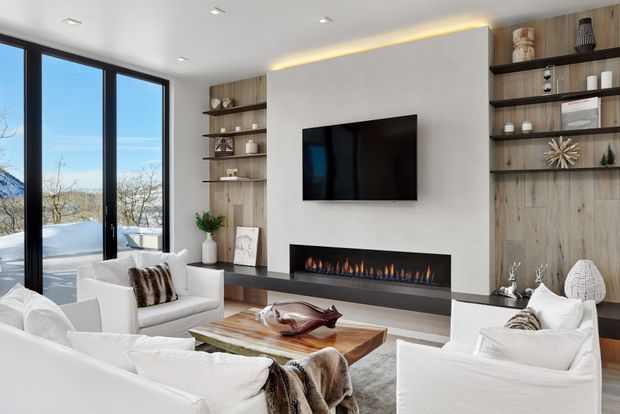 Contemporary living room with floor-to-ceiling black-framed glazing, a wall-mounted television, long linear fireplace, and bespoke open shelving integrated into warm timber wall panelling. White upholstered seating surrounds a substantial timber coffee table, while concealed LED lighting adds a refined glow above the feature wall. Snow-covered landscape is visible outside.