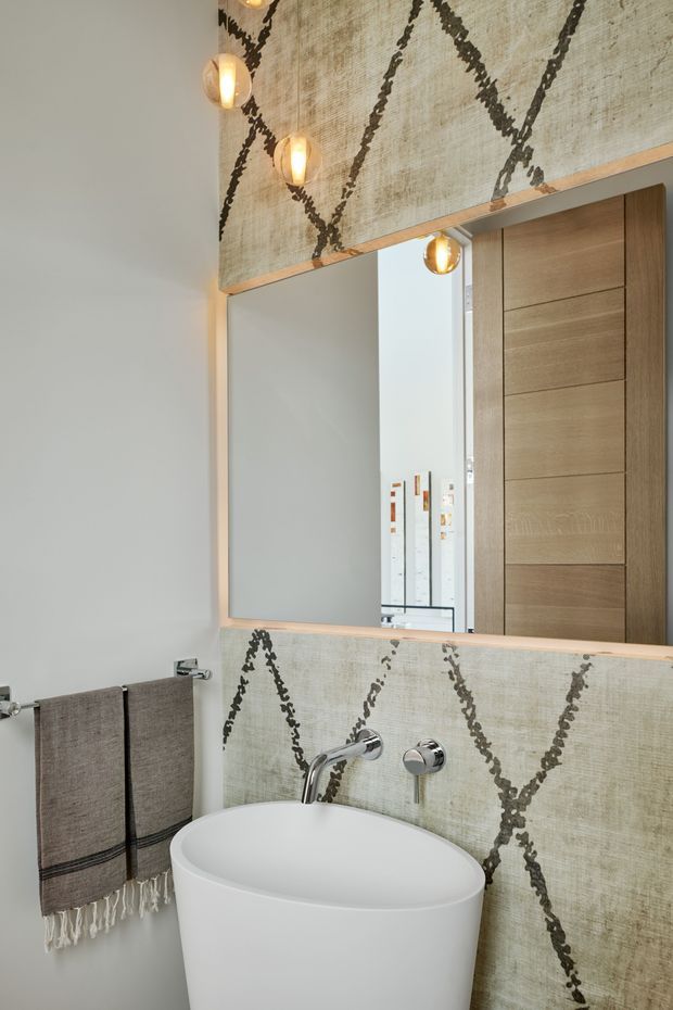 A compact contemporary bathroom with a freestanding white vessel basin, wall-mounted chrome tapware, a large timber-framed mirror, and a timber door reflected in the mirror. The wall behind the basin and above the mirror features a textured beige surface with bold dark geometric lines. Warm globe pendant lights hang beside and above the mirror, while dark taupe fringed towels add contrast.