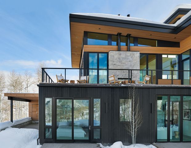 Exterior view of a contemporary multi-level house in a snowy setting. The building features a flat, layered roofline, dark vertical cladding on the lower level, warm timber soffits and upper wall cladding, expansive black-framed glazing, and a raised terrace with slender metal balustrades. A central stone-clad wall provides a contrasting feature surface.