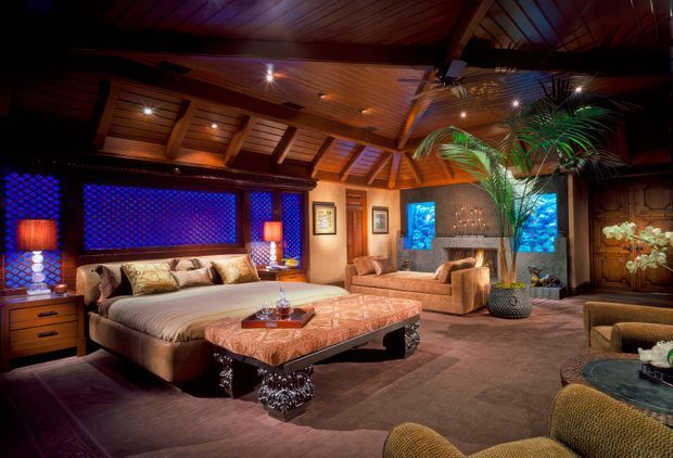 A richly styled luxury bedroom with a large upholstered bed, matching bedside tables and lamps, a carved upholstered bench, seating area, dark timber walls and a dramatic exposed timber-beam ceiling. Blue patterned window screens, tropical palms, ornate furnishings and warm ambient lighting create an exotic, opulent atmosphere.