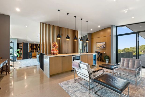 Open-plan contemporary kitchen and living area with a large timber island, vertical timber screen, black pendant lights, and integrated cabinetry. The foreground contains a relaxed seating arrangement with leather chairs and ottoman on a patterned rug. Full-height glazing opens to an outdoor terrace and leafy outlook, while built-in shelving and additional living space are visible beyond the kitchen.