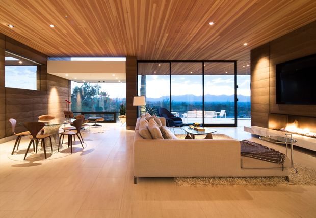 Open-plan contemporary living and dining area with expansive floor-to-ceiling glazing overlooking a mountain landscape. The room features a warm timber slatted ceiling, timber wall cladding, pale large-format flooring, a beige sectional sofa, dining table and chairs, recessed lighting, and a dark feature wall with a large television and linear fireplace.