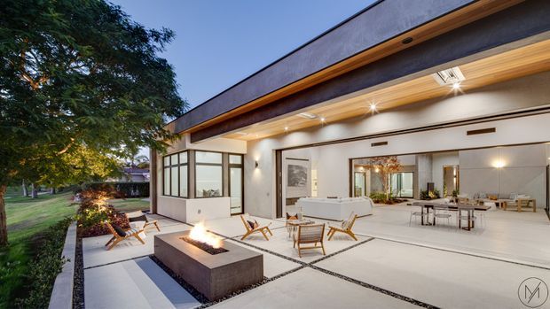 Twilight exterior view of a contemporary single-storey residence opening onto a large paved entertaining terrace. The image features broad retractable or sliding glazed openings, a timber-lined deep eave, pale rendered walls, a flat roofline, and a sunken square fire pit surrounded by outdoor lounge chairs. The open interior is visible through the glazing, including a living area and dining space. Mature landscaping borders the terrace.