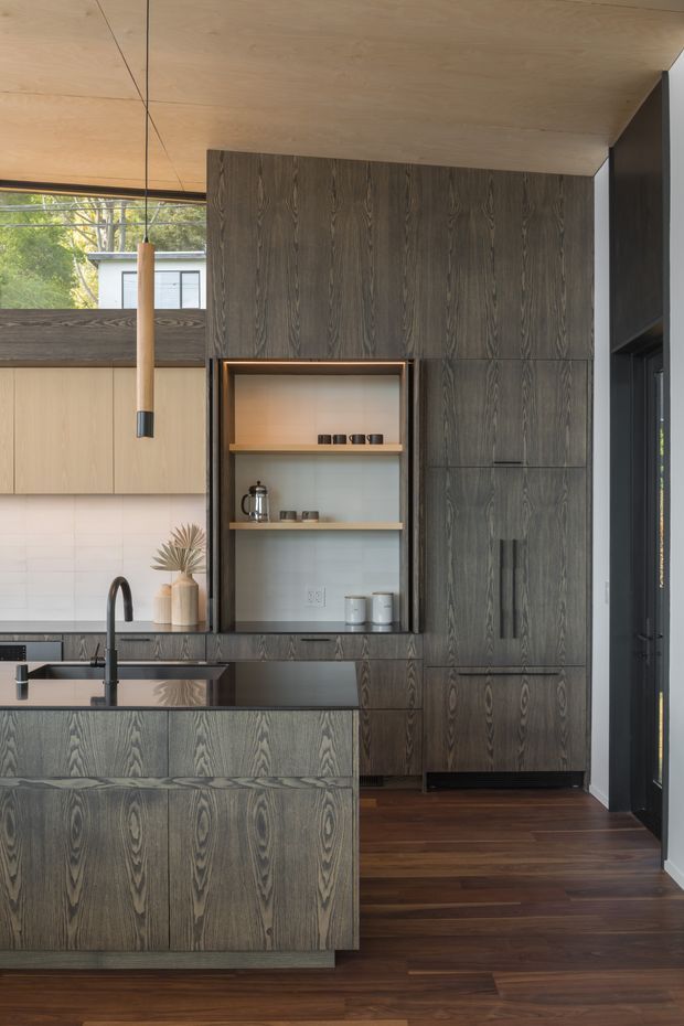 A refined contemporary kitchen with a large island, integrated sink and dark gooseneck tap, extensive dark timber cabinetry, open display shelving, and a contrasting light timber cabinetry section. The kitchen features a warm timber ceiling, pale tiled splashback, dark stone or composite benchtops, and rich dark timber flooring. Full-height glazing and a glazed door bring in natural light and greenery from outside.
