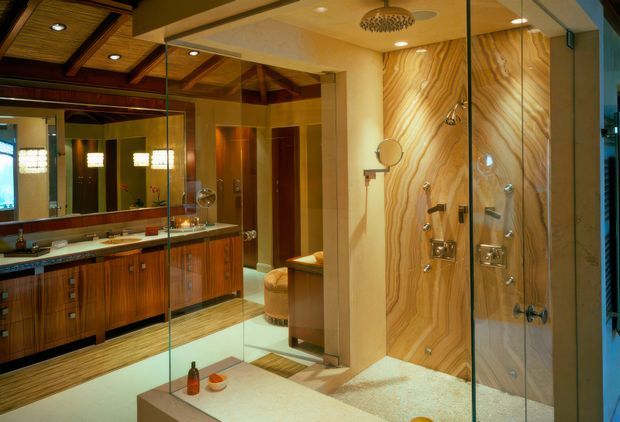 A luxurious bathroom with a large frameless glass shower enclosure, dual vanity, and extensive mirrored surfaces. The space features warm timber cabinetry, a dramatic flowing-grain wood feature wall inside the shower, a timber-lined coffered ceiling with exposed beams, polished stone-look benchtops, and warm integrated lighting. A freestanding or partially visible bath and upholstered stool add to the refined, spa-like atmosphere.