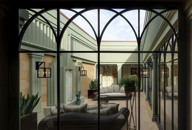 View through a dark metal-framed glazed wall into a refined enclosed courtyard. The courtyard features pale green and stone-toned walls, repeated arched glazing and openings, black decorative lanterns, tiled paving, potted plants, and woven outdoor-style seating arranged around a central table. The image is architectural and interior-facing, with strong symmetry and a calm, elegant atmosphere.