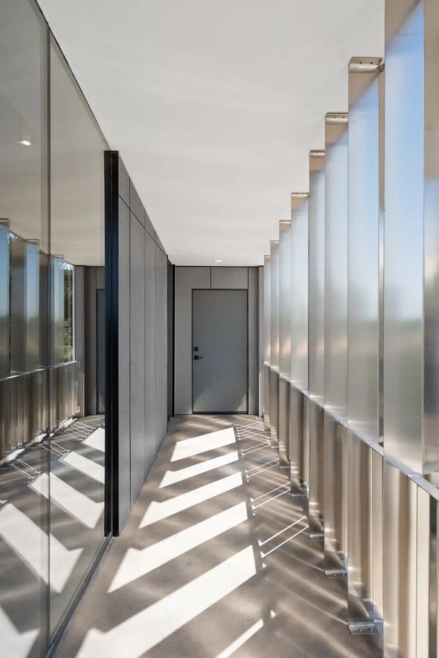A long, narrow contemporary interior corridor with a polished concrete floor, full-height glazing and reflective surfaces on the left, and a rhythmic row of translucent or light-coloured vertical panels with metal supports on the right. A dark grey flush door terminates the passage. Strong sunlight creates bold diagonal geometric shadows across the floor and walls, producing a bright, minimalist architectural atmosphere.