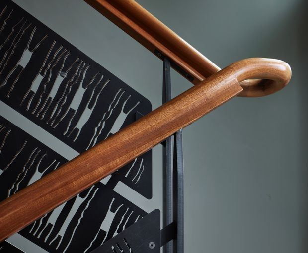 Close-up architectural detail of a polished timber stair handrail with a curved end, mounted beside dark metal framing and decorative perforated metal balustrade panels. The surrounding wall and ceiling are painted in a muted blue-grey tone. The image emphasizes craftsmanship, joinery, and the contrast between warm wood and dark metal.