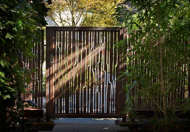 A secluded garden entry framed by dense greenery, featuring a tall dark timber slatted gate and matching side fencing. Sunlight filters through the narrow vertical gaps, creating dappled shadows across the gate and paved path.