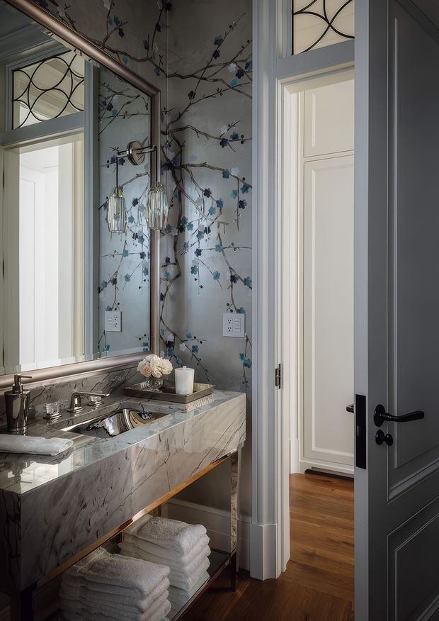 A refined powder room viewed from the doorway, featuring a wall-mounted marble vanity with an integrated basin, polished metal tapware, open towel storage below, and a large framed mirror. The walls are covered in a botanical mural or patterned wallpaper with blue flowers and branches, complemented by decorative frosted-glass transom panels and dark paneled doors with black hardware. Warm timber flooring is visible through the adjoining doorway.