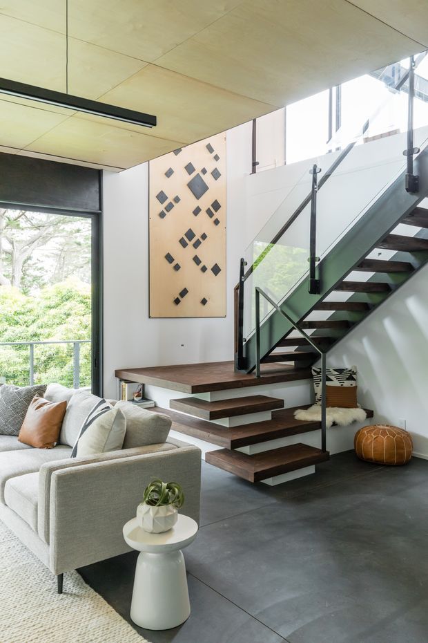 Bright contemporary open-plan living area featuring a floating timber staircase with dark wood treads, black metal supports, and clear glass balustrades. The room has a smooth dark grey floor, light walls, a pale timber-paneled ceiling, large glazing overlooking greenery, and a neutral upholstered sofa. A geometric wall artwork adds a small decorative accent.