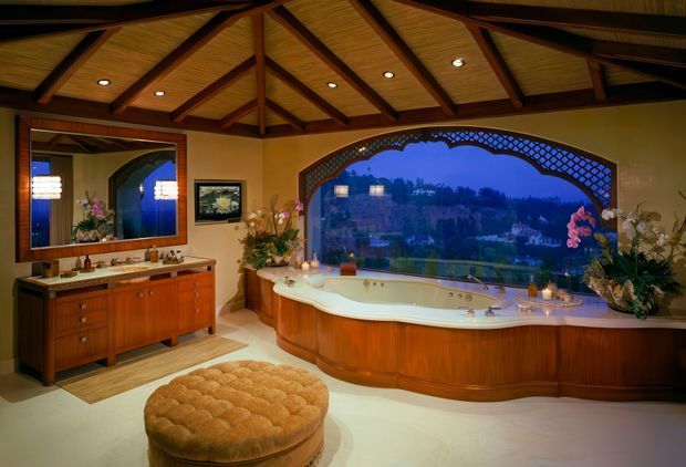 Luxurious bathroom with a large curved built-in spa bath positioned beside an expansive arched picture window overlooking a distant landscape. The room features warm timber cabinetry, a large framed mirror, decorative plants, cream walls, and a dramatic exposed timber vaulted ceiling with dark beams and recessed lighting.