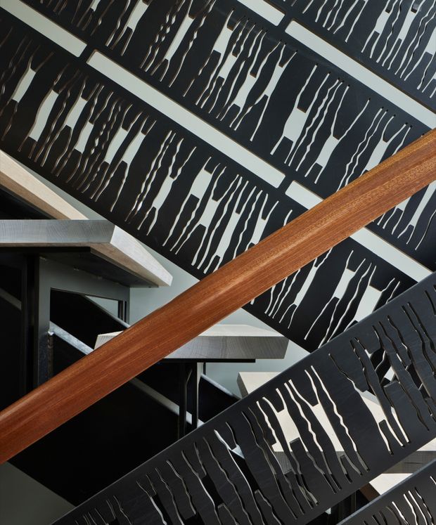 Close-up architectural view of a sculptural stairway with multiple dark metal balustrade panels featuring irregular vertical cut-outs. Bold timber handrails cross the composition, with exposed concrete or light-toned structural elements and strong geometric diagonals. The space has a dramatic, contemporary industrial character.