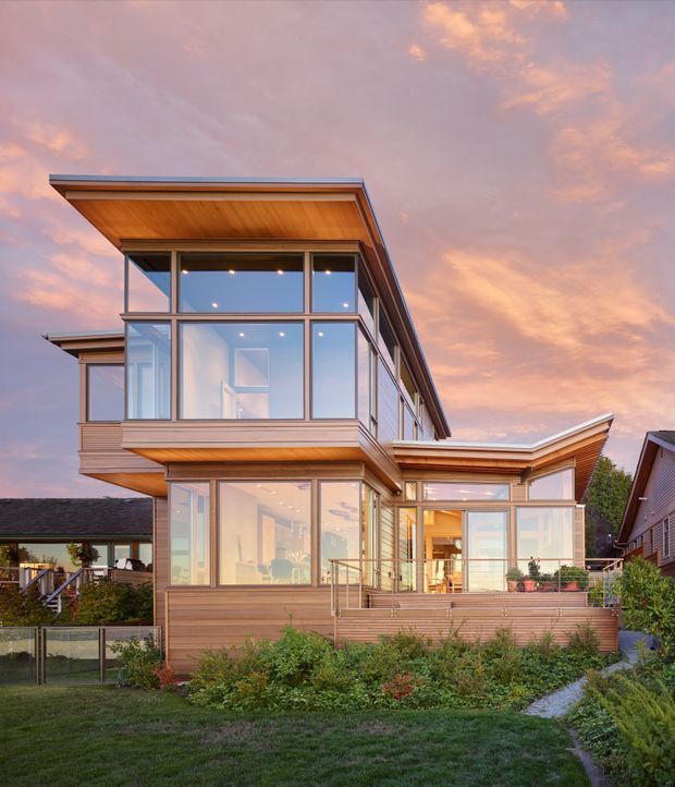 Exterior view of a contemporary multi-level home with extensive floor-to-ceiling glazing, warm timber soffits and horizontal timber-clad walls. The building has projecting upper volumes, angular rooflines, balconies or terraces, and strong indoor-outdoor visual connection. It is set in a landscaped suburban garden and photographed at dusk beneath a pink-orange sky.