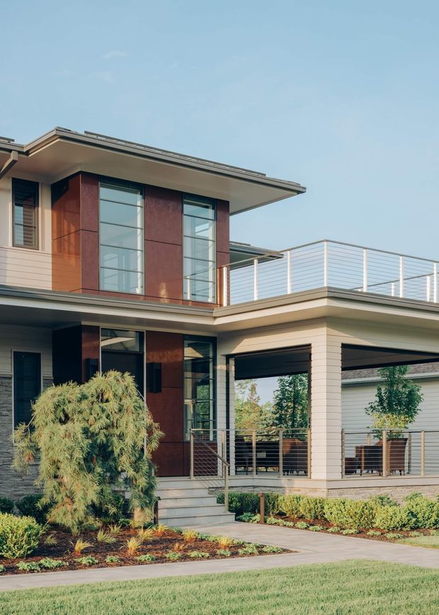 Exterior view of a contemporary multi-level house with a low-pitched roof, broad roof overhangs, large vertical glazed windows, warm reddish-brown facade panels, horizontal light-coloured cladding, and a covered open carport or outdoor parking area with a rooftop terrace and cable-style railing. Landscaped planting beds, paving, and lawn occupy the foreground.