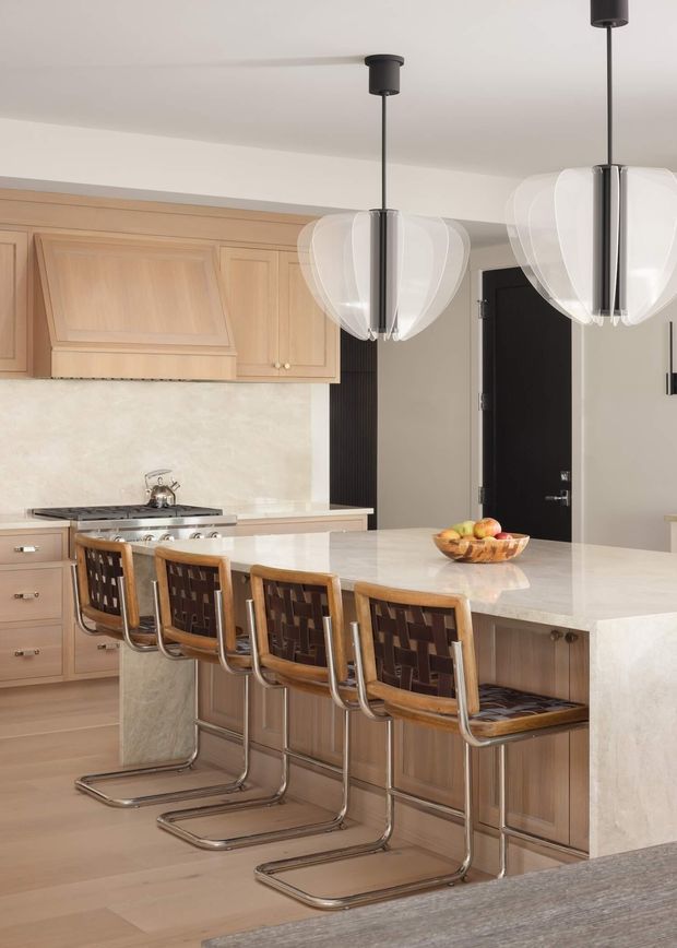A refined contemporary kitchen centered on a large island with a pale stone benchtop and integrated seating for five. The kitchen features warm light-wood framed cabinetry, a matching stone splashback, stainless-steel appliances, and distinctive oversized translucent pendant lights with dark metal stems. A dark internal door and vertical black wall detail provide contrast, while the overall palette is warm and neutral.