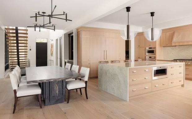 Bright open-plan kitchen and dining area with pale timber cabinetry, a large pale stone island, light timber flooring, a white dining table, upholstered dining chairs, and sculptural black linear pendant lighting. A dark entry door and timber slatted window screening are visible in the background.