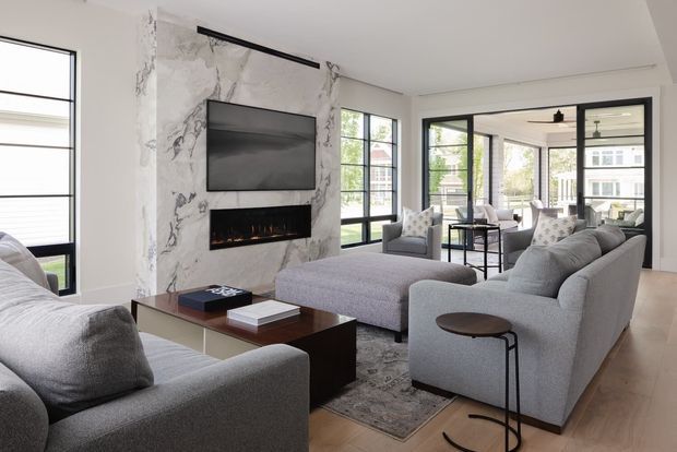 Bright contemporary living room with a large grey sectional sofa and matching armchair arranged around a dark wood coffee table. A marble-look feature wall incorporates a wall-mounted television and linear fireplace, while black-framed glazing opens the room toward a sunroom or enclosed terrace. Pale timber flooring, neutral upholstery, and minimal black accents create a refined, airy interior.