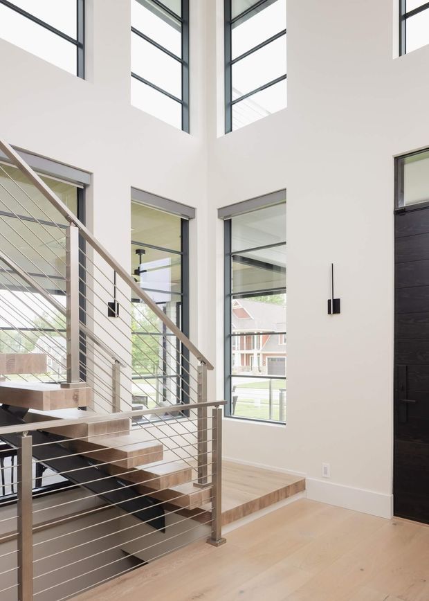 Bright contemporary residential entryway with a floating timber staircase, slim horizontal metal balustrade, pale timber flooring, tall black-framed windows, double-height glazing, and a dark pivot front door. The space is minimalist and airy, with strong vertical volume and abundant natural light.