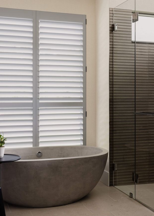 A refined contemporary bathroom featuring a large freestanding concrete bathtub beside a frameless glass shower enclosure. White plantation shutters provide strong daylight, while the shower wall is finished in dark horizontally textured tiles. The space has a neutral palette of concrete grey, white, and dark grey with understated modern fixtures.