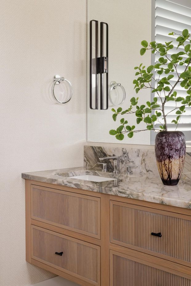 Close-up of an elegant bathroom vanity with a double-basin marble countertop and matching marble splashback. The vanity features warm fluted timber cabinetry with dark pulls, while a large mirror, chrome towel rings and tapware, a sculptural black-and-metal wall light, and a decorative plant complete the refined fit-out.