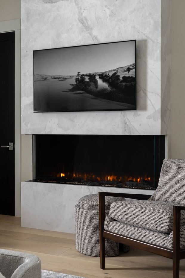 A refined contemporary living room vignette featuring a large linear fireplace set into a pale veined stone surround, with a wall-mounted television above. A textured upholstered lounge chair and matching ottoman sit on light timber flooring beside the fireplace. The palette is restrained in white, grey, black and natural wood, with warm firelight providing contrast.