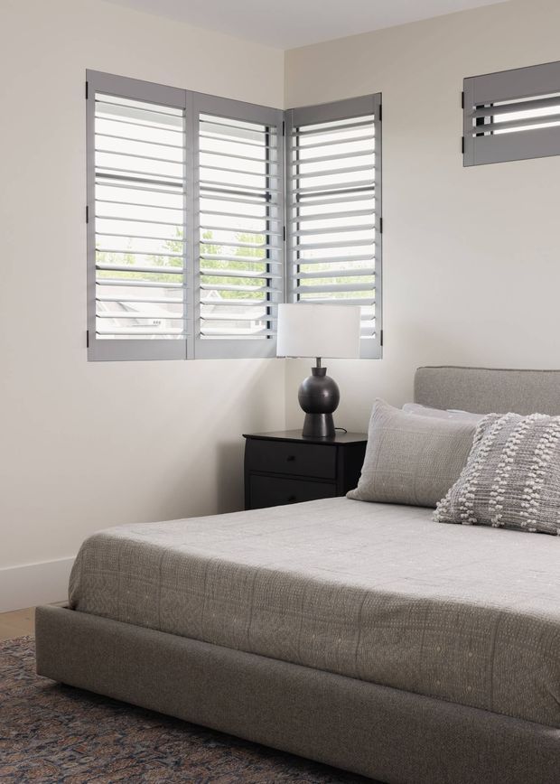 A softly lit contemporary bedroom with a large upholstered bed in a pale grey textured fabric, layered neutral bedding and patterned cushions, and a dark bedside chest with a rounded black table lamp. Grey plantation shutters cover the corner windows, with matching horizontal shutter panels on the adjacent wall. The palette is neutral and muted, with a patterned area rug partially visible beneath the bed.
